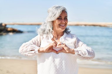 Middle age woman smiling confident doing heart gesture with hands at seaside