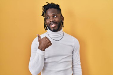 Sarı arka planda balıkçı yaka kazak giyen Afrikalı bir adam gülümsüyor ve başparmağıyla yana bakıyor.. 