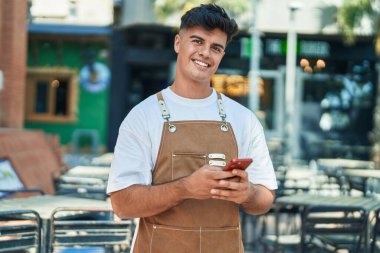 Genç İspanyol garson, kahvecinin terasında akıllı telefon kullanarak gülümsüyor.