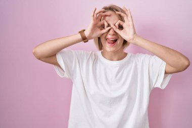Pembe arka planda duran genç, beyaz bir kadın dürbün gibi jest yapıyor. Dürbün gibi dilini çıkarıyor, gözler parmaklarından bakıyor. çılgın ifade. 