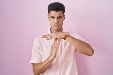 Young hispanic man standing over pink background doing time out gesture with hands, frustrated and serious face 