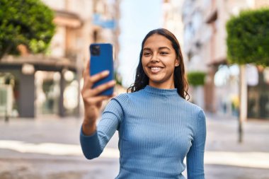 Genç Afrikalı Amerikalı kadın gülümsüyor. Kendine güveni tam. Sokakta akıllı telefondan selfie çekiyor.