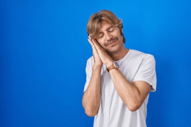 Orta yaşlı bir adam mavi arka planda ayakta uyur, yorgun düşler kurar ve elleri ile poz verir, gözleri kapalı gülümserdi.. 