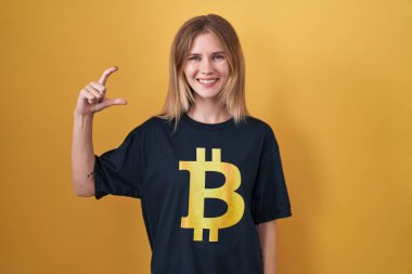 Sarışın, beyaz kadın bitcoin tişörtü giyiyor gülümsüyor ve kendine güvenen el hareketleriyle küçük beden işareti yapıyor parmaklarıyla bakıyor ve kamera var. ölçüm kavramı. 