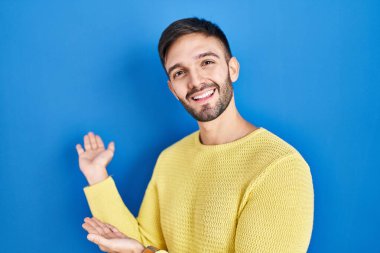 Hispanic man standing over blue background inviting to enter smiling natural with open hand 