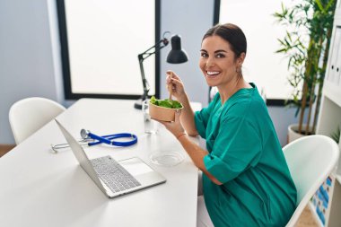 Genç, güzel İspanyol kadın doktor. Klinikte salata yerken kendinden emin gülümsüyor.