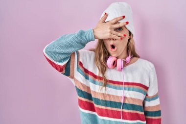 Young blonde woman standing over pink background peeking in shock covering face and eyes with hand, looking through fingers with embarrassed expression. 