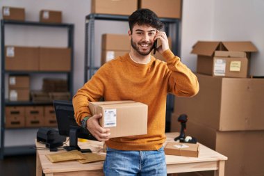 Young hispanic man ecommerce business worker talking on smartphone holding package at office