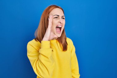Young woman standing over blue background shouting and screaming loud to side with hand on mouth. communication concept. 