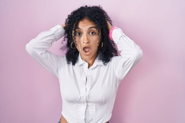 Pembe arka planda duran kıvırcık saçlı İspanyol kadın çıldırmış ve elleri başının üstünde korkmuş, ağzı açık şoktan korkmuş ve şaşırmış. 