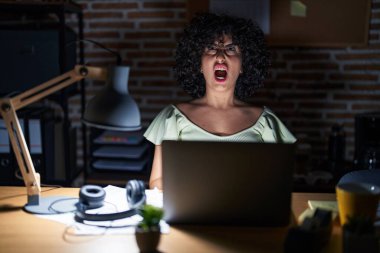 Young brunette woman with curly hair working at the office at night angry and mad screaming frustrated and furious, shouting with anger. rage and aggressive concept. 