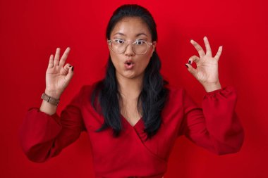 Asyalı genç bir kadın kırmızı arka planda duruyor. Şaşırmış ve şok olmuş görünüyor. Tamam sembolünü parmaklarıyla yapıyor. çılgın ifade 