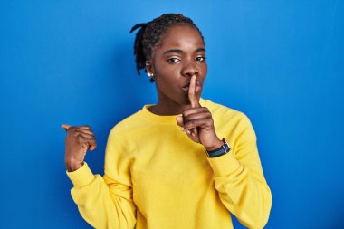 Güzel siyahi kadın mavi arka planda dikiliyor, sessiz olmak istiyor, parmakları dudaklarında, elleri yana doğru işaret ederek. sessizlik ve gizli konsept. 