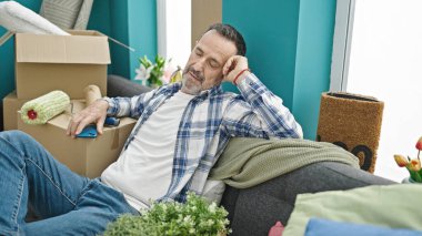 Orta yaşlı bir adam elinde akıllı bir telefonla kanepeye oturmuş yeni evinde uyuyor.