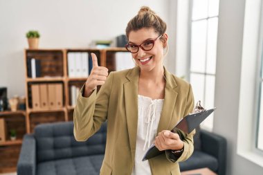 Muayenehanede çalışan genç bir kadın elleriyle mutlu bir jest yapıyor. Kameraya bakıp başarıyı gösteren ifade. 
