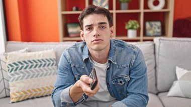 Young hispanic man watching tv sitting on sofa with boring expression at home