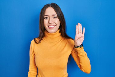 Genç esmer kadın mavi arka planda duruyor ve parmaklarını dört numarayı gösterip işaret ederken kendinden emin ve mutlu bir şekilde gülümsüyor.. 