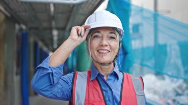 Orta yaşlı, gri saçlı kadın müteahhit sokakta kendine güvenerek gülümsüyor.