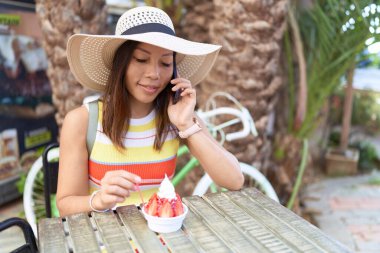 Genç Asyalı turist dondurma yiyor. Kahve dükkanının terasında akıllı telefondan konuşuyor.