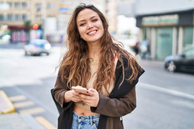 Genç, güzel, İspanyol bir kadın sokakta akıllı telefon kullanarak gülümsüyor.