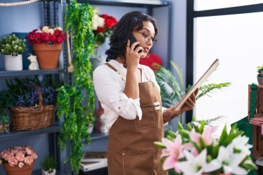 Young beautiful hispanic woman florist talking on smartphone reading clipboard at flower shop