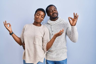 Mavi arka planda birlikte duran genç Afro-Amerikan çift rahatlamış ve gözleri kapalı parmaklarıyla meditasyon hareketi yaparken gülümsüyor. yoga konsepti. 