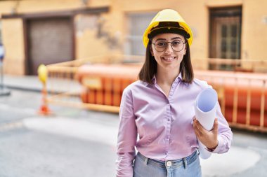 Hispanic girl wearing architect hardhat at construction site looking positive and happy standing and smiling with a confident smile showing teeth 
