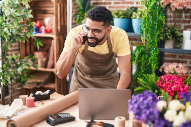 Young arab man florist talking on smartphone using laptop at florist