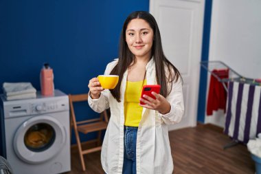 Genç İspanyol kadın akıllı telefon kullanıyor ve kahve içiyor. Çamaşır odasında çamaşır makinesini bekliyor.