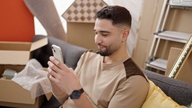 Genç Arap adam yeni evinde akıllı telefon kullanarak kendinden emin gülümsüyor.