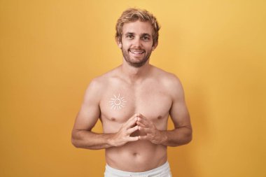 Beyaz adam güneş kremi sürmüş üstsüz duruyor ve parmaklarını çapraz yapıp gülümsüyor rahat ve neşeli. Başarı ve iyimserlik 