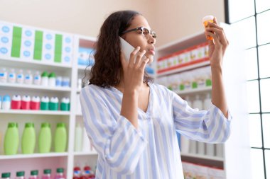 Young african american woman customer talking on smartphone holding pills bottle at pharmacy