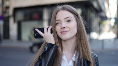 Young blonde woman smiling confident listening audio message by the smartphone at street