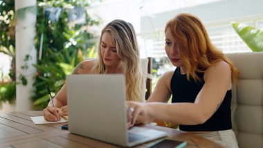 Ev terasındaki masada dizüstü bilgisayarda yazan iki kadın.