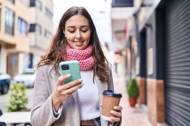 Young beautiful hispanic woman using smartphone drinking coffee at street