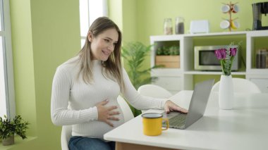Genç hamile kadın yemek odasındaki masada video görüşmesi yapıyor.