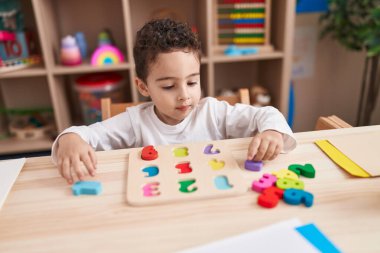 Adorable hispanic boy playing with maths puzzle game sitting on table at kindergarten