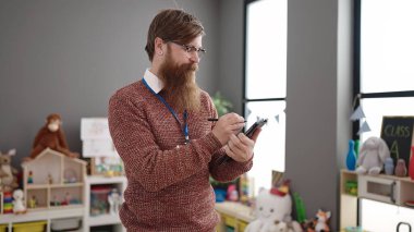Young redhead man preschool teacher writing on touchpad standing at kindergarten