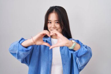 Beyaz arka planda duran Çinli genç bir kadın. Sevgiyle gülümsüyor. Elleriyle kalp sembolü yapıyor. romantik kavram. 