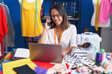 Young hispanic woman tailor using laptop talking on smartphone at sewing studio