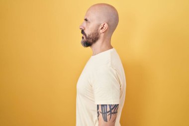 Sarı arka planda dikilen İspanyol dövmeli adam yan bakıyor, rahat poz veriyor ve kendine güvenen bir gülümseme takınıyor.. 