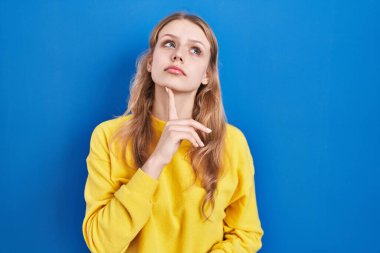 Young caucasian woman standing over blue background thinking concentrated about doubt with finger on chin and looking up wondering 