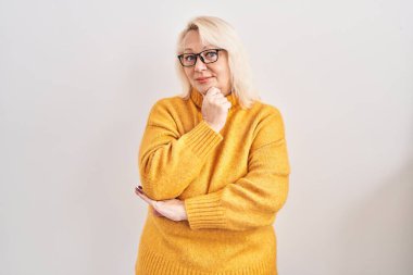 Orta yaşlı beyaz kadın arka planda duran gözlüklü kameraya kendinden emin bir şekilde bakıyor kollarını kavuşturmuş ve çenesini kaldırmış bir şekilde gülümsüyor. Olumlu düşünmek. 