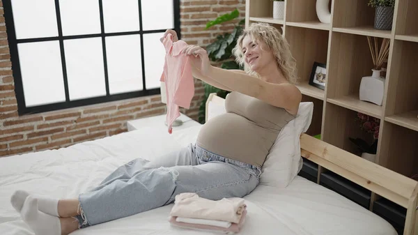 Young Pregnant Woman Holding Baby Clothes Sitting Bed Bedroom — стоковое фото