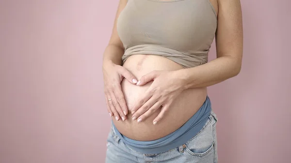 若いです妊娠中の女性あります心ジェスチャーとともに手上の腹上の隔離されたピンクの背景 — ストック写真