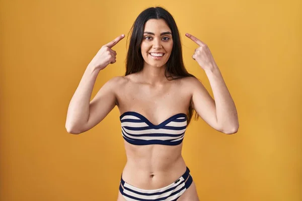 Mujer Morena Joven Con Bikini Sobre Fondo Amarillo Sonriendo Apuntando — Foto de Stock