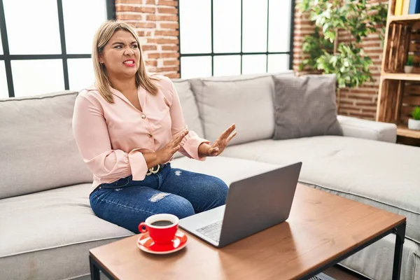 Young hispanic woman using laptop sitting on the sofa at home disgusted expression, displeased and fearful doing disgust face because aversion reaction. with hands raised