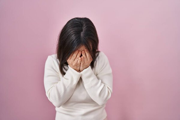 Woman with down syndrome standing over pink background with sad expression covering face with hands while crying. depression concept. 