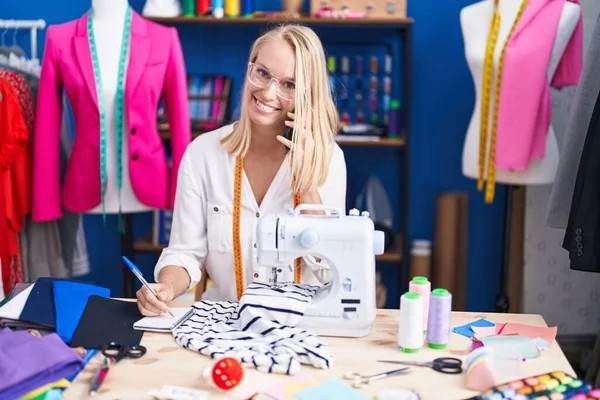 Young Blonde Woman Tailor Talking Smartphone Writing Notebook Sewing Studio — Stockfoto