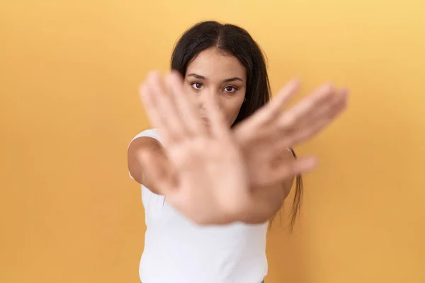 Young arab woman wearing casual white t shirt over yellow background rejection expression crossing arms and palms doing negative sign, angry face